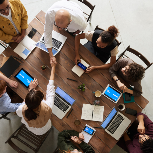 An overhead view of a group of diverse professionals seated around a large wooden table, engaged in a collaborative meeting. The table is scattered with laptops, notebooks, smartphones, and coffee cups. Two individuals in the center are shaking hands, symbolizing agreement or teamwork, while others interact and take notes. The setting reflects a productive and cooperative work environment.